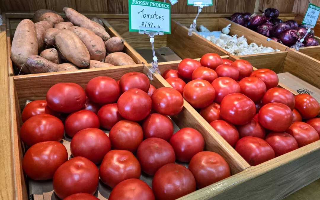 Family Foods Edina MO Wooden crates filled with fresh vegetables, including red tomatoes priced at $1.99/lb and other produce like sweet potatoes, onions, and garlic at a market.