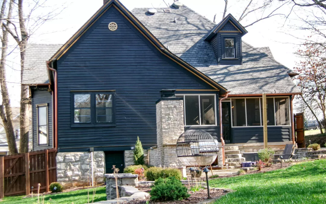 Miller's Guttering Munfordville, KY A two-story dark blue house with new gutters, stone accents, and a screened porch, surrounded by a landscaped garden and trees under a clear sky.