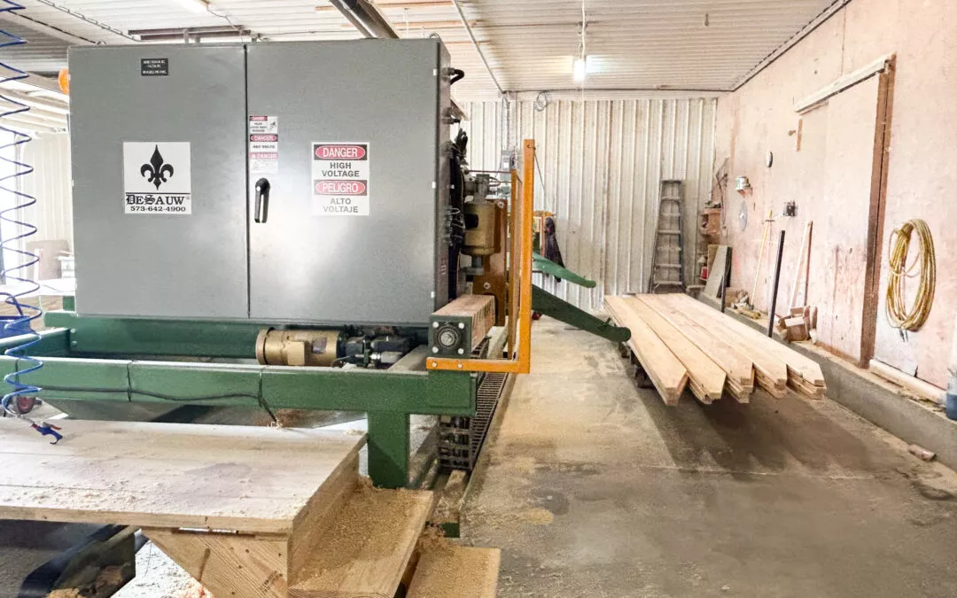 Martin's Truss Manufacturing Elkhart IN A large industrial saw and conveyor belt in a wood processing facility, with stacks of lumber and exposed interior walls.