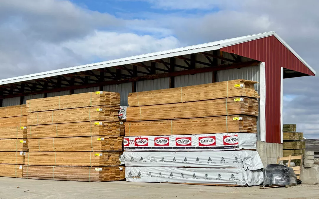 Martin's Truss Manufacturing Elkhart IN A large outdoor warehouse storing stacks of lumber under a corrugated metal roof, with some stacks covered in branded protective wraps.