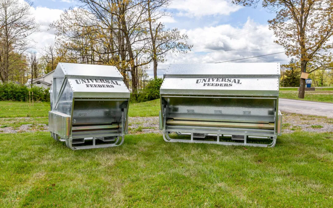 Schmucker's Livestock Handling Horse Cave, Kentucky Two large metal bulk feeders labeled "universal feeders" positioned on a grassy area beside a road under a cloudy sky.