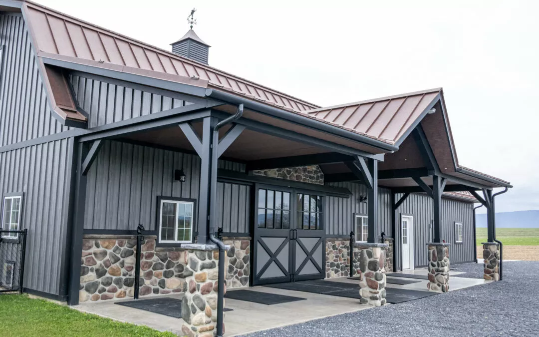 Martin's Trim Forming Millmont, PA A gray barn with a metal roof, a weather vane on top, wooden beams, and a stone base stands on a paved area with a grassy yard nearby.