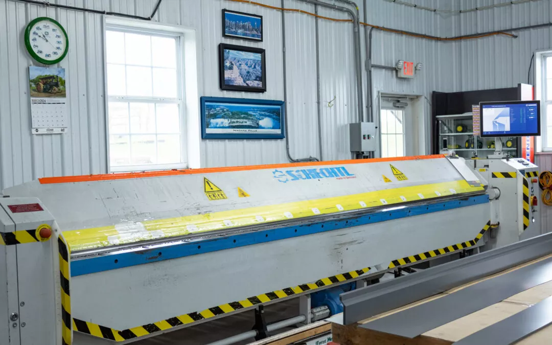 Martin's Trim Forming Millmont, PA A sheet metal bending machine in an industrial room, with safety warnings. The room has a wall clock, framed pictures, and a computer screen showing digital controls.