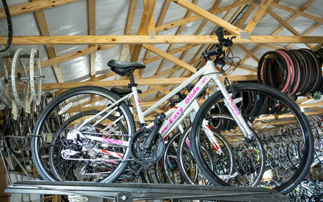 H & H Bicycles Liberty Kentucky A white bicycle with "Easy Glide" written on the frame is displayed on a rack in a wooden-roofed bike shop, surrounded by other bicycles and spare parts.