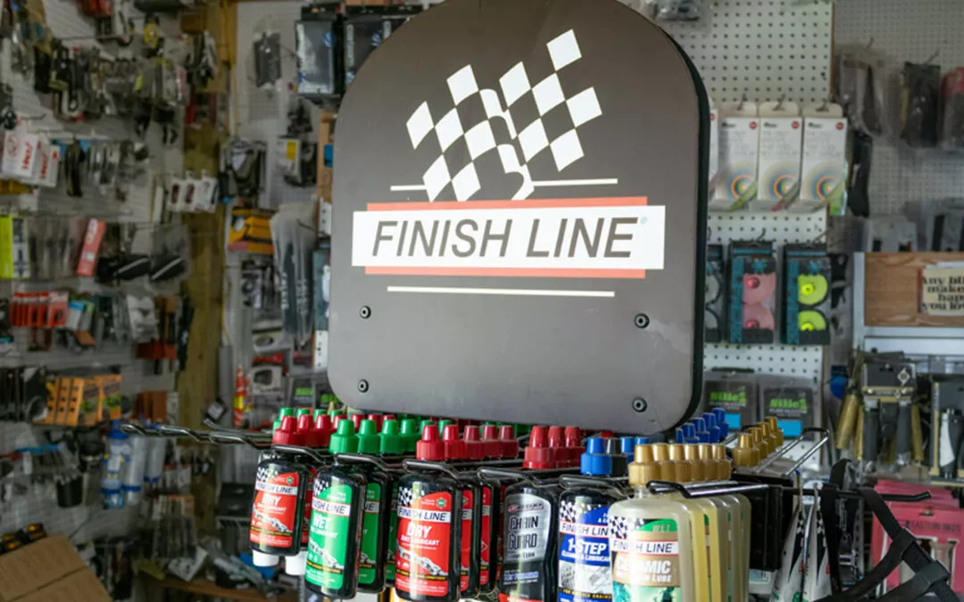 H & H Bicycles Liberty Kentucky Display of various bottles of Finish Line bicycle lubricants on a shelf in a store, with a Finish Line sign featuring a checkered flag logo above them. Tools and accessories are visible in the background.