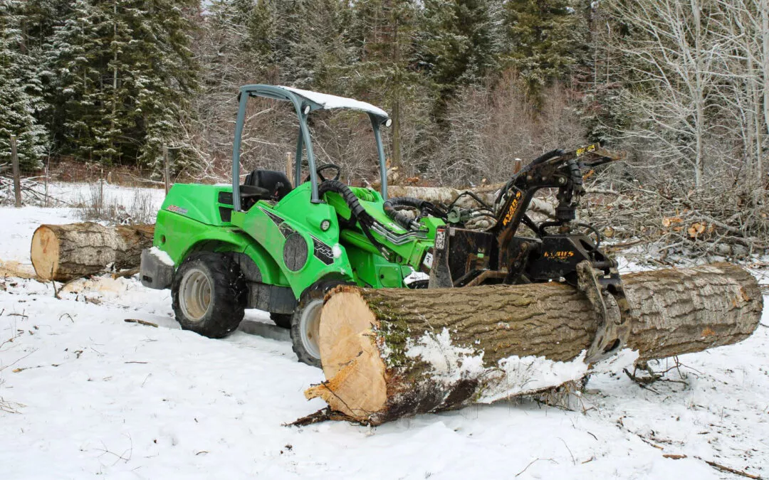 6.24.24 Treeco Gallery 1 A green tractor equipped with a log grapple attachment is lifting a large tree log in a snowy forested area.