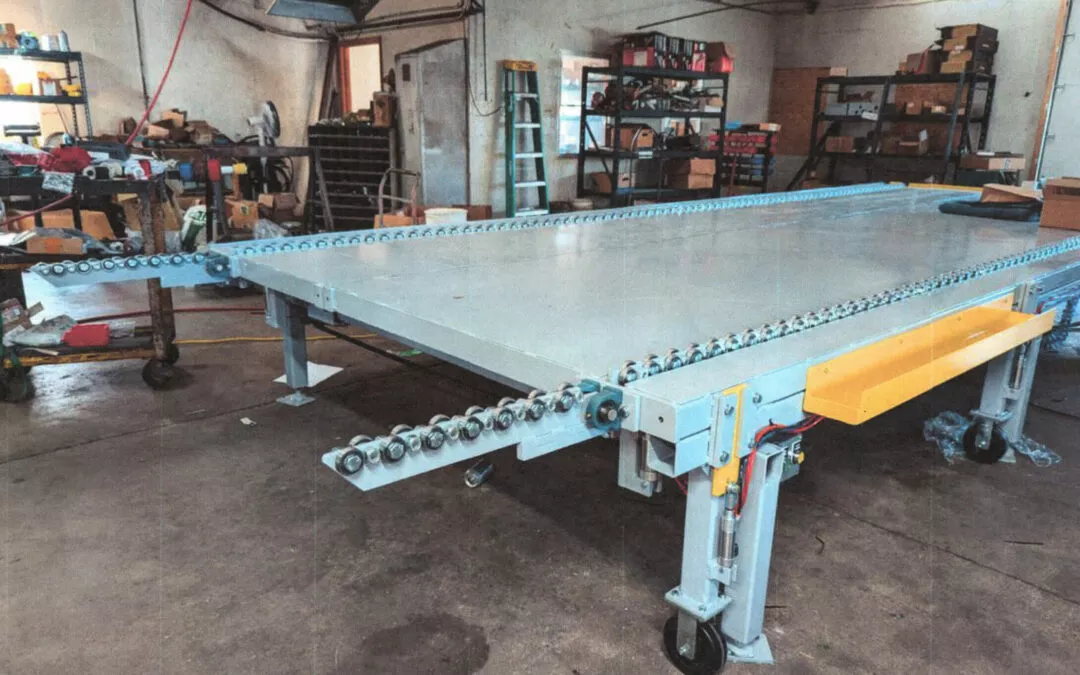 7.19.24 MidState MMidstate Lamp LLC Arthur Illinoisachine Gallery 1 A large industrial metal conveyor assembly with rollers and a yellow safety barrier is stationed in a workshop filled with various tools, shelves, and machinery.