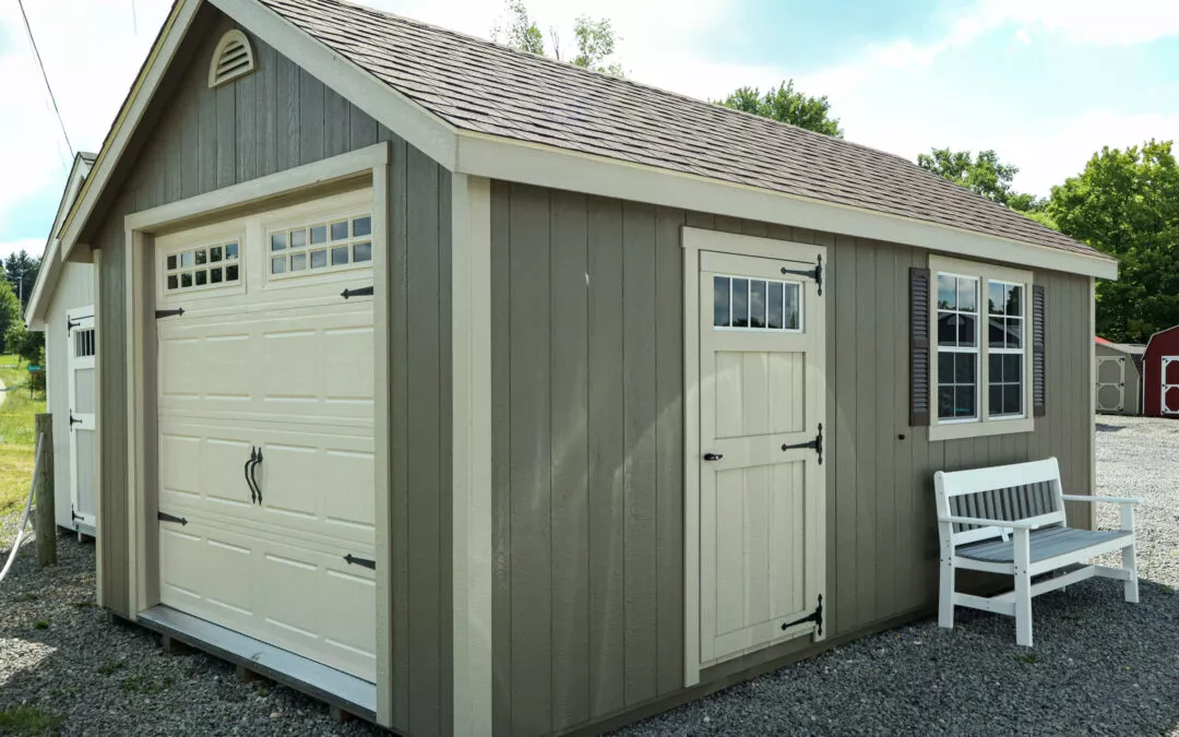 Summit Structures & Outdoor Living Greenville, PA A small, gray storage shed with a white garage door, a white entrance door, and two windows. A white bench is placed beside the entrance door. Gravel ground surrounds the shed.