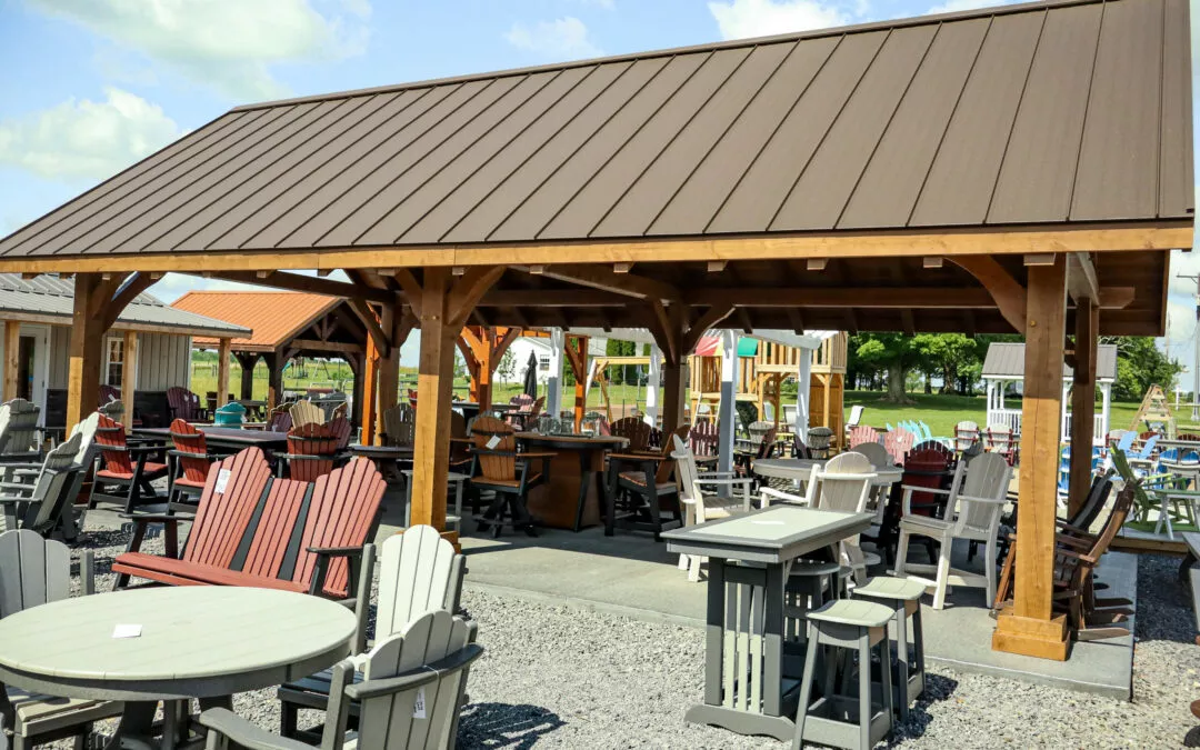 Summit Structures & Outdoor Living Greenville, PA Outdoor showroom displaying a variety of wooden patio furniture, including chairs, tables, and benches, under and around a wooden pavilion with a metal roof on a sunny day.