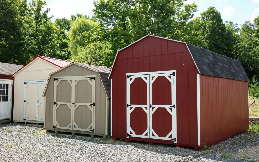 Summit Structures & Outdoor Living Greenville, PA Three storage sheds in red, beige, and cream colors are placed on a gravel area, with a background of green trees and a partly cloudy sky.