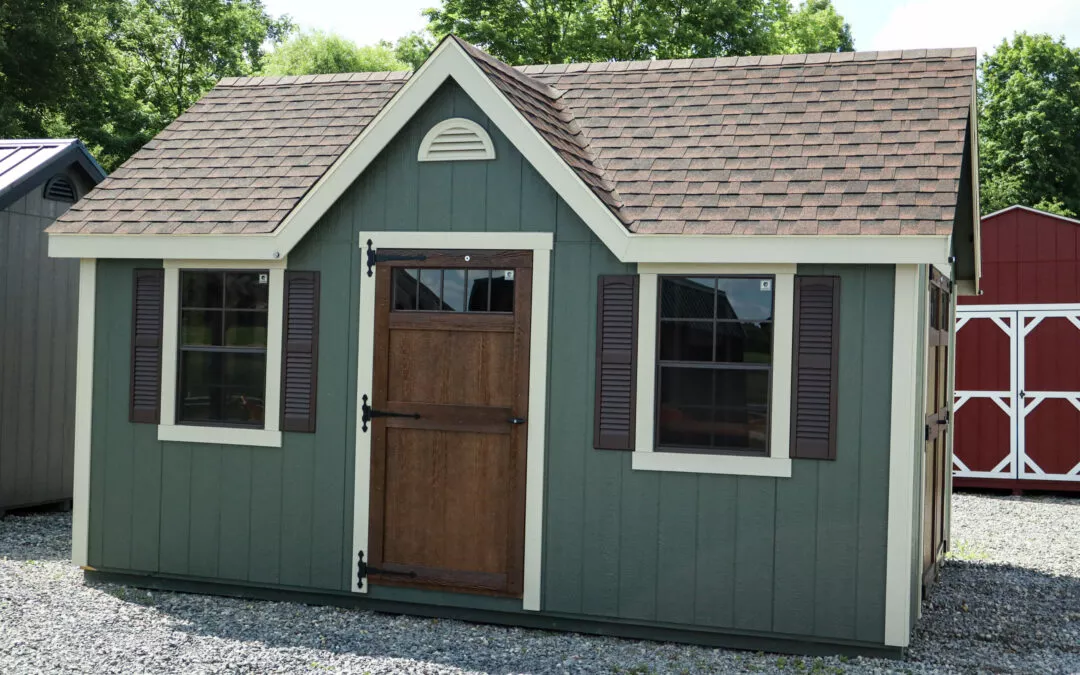 Summit Structures & Outdoor Living Greenville, PA Small green shed with brown shutters and a matching brown door, featuring two windows on each side of the door and a gabled roof. The shed stands on a gravel surface with trees in the background.