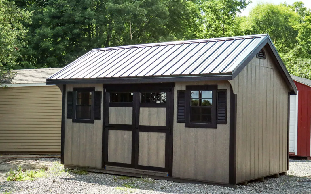 Summit Structures & Outdoor Living Greenville, PA A small, tan shed with a dark roof and black trim is situated on a gravel area, surrounded by trees and other outbuildings.