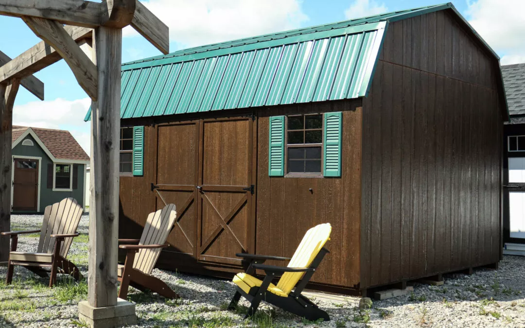 Summit Structures & Outdoor Living Greenville, PA A brown shed with green shutters and a green metal roof, surrounded by lawn chairs, sits on a gravel area under a clear sky.