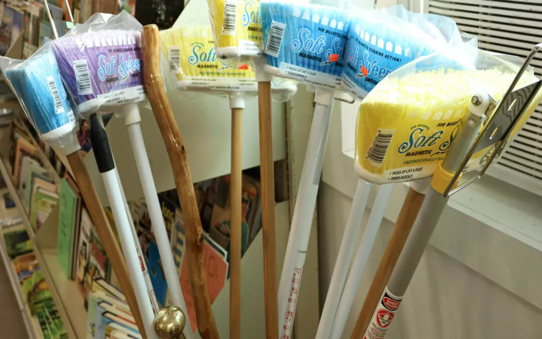 Countrty Pantry Sheridan, MI Several cleaning brushes with various colored bristles and labels are displayed standing upright in a store, alongside bookshelves filled with books.