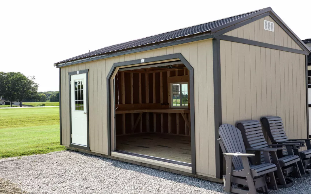 Cedar Ridge Sales Ramsey IL Ava IL A small, beige storage shed with an open door is seen on a gravel lot. Several dark gray Adirondack chairs are stacked beside the shed. A grassy field and trees are visible in the background.