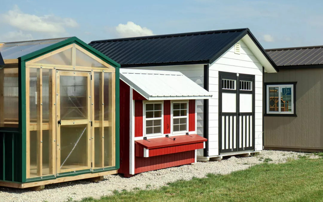 Cedar Ridge Sales Ramsey IL Ava IL Array of small shed buildings including a green greenhouse, a red and white playhouse, and a white shed with black trim, situated on gravel with a grassy area in the foreground.
