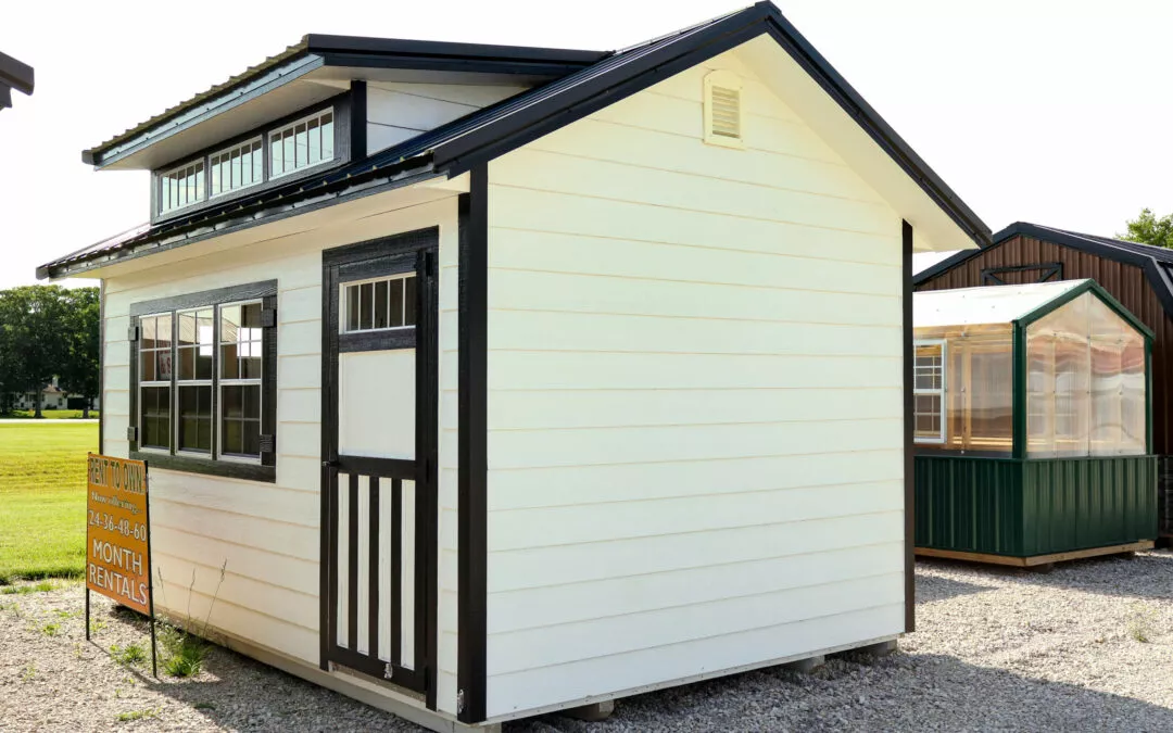 Cedar Ridge Sales Ramsey IL Ava IL A small, white, single-story shed with black trim and a sign advertising month rentals in front. Another shed, green and transparent, is partially visible in the background.