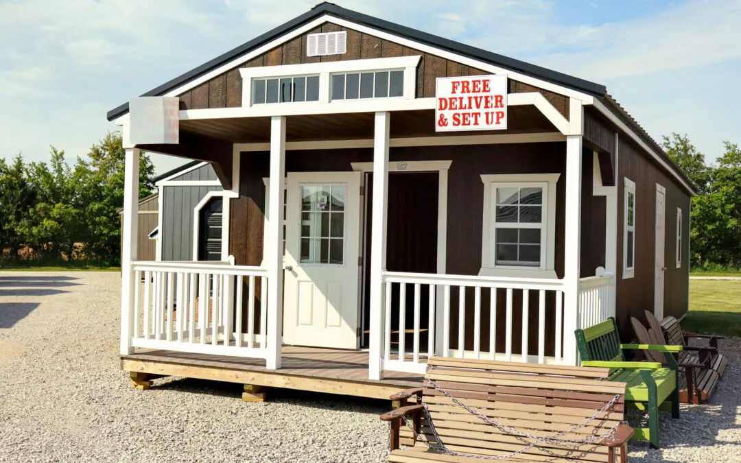 Cedar Ridge Sales Ramsey IL Ava IL A small brown wood cabin with a porch and a "Free Delivery & Set Up" sign. It is situated on a gravel area with some green chairs and benches in front.