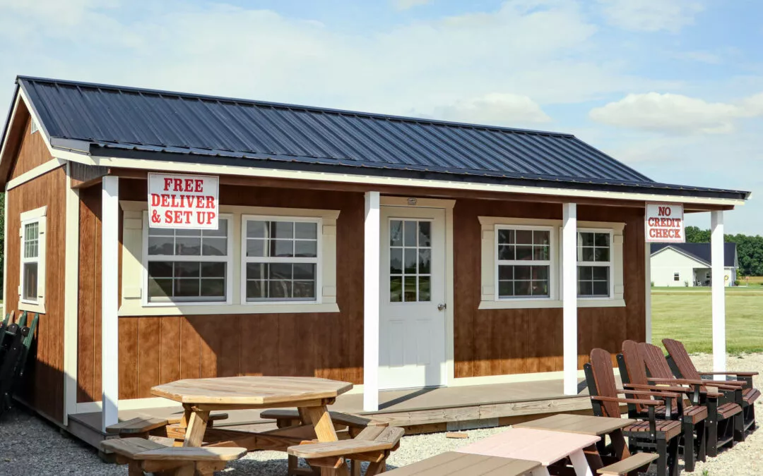 Cedar Ridge Sales Ramsey IL Ava IL A small brown and white building with a dark roof, signs saying "Free Delivery & Set Up" and "No Credit Check," benches and picnic tables outside on a gravel area, and green lawn in the background.