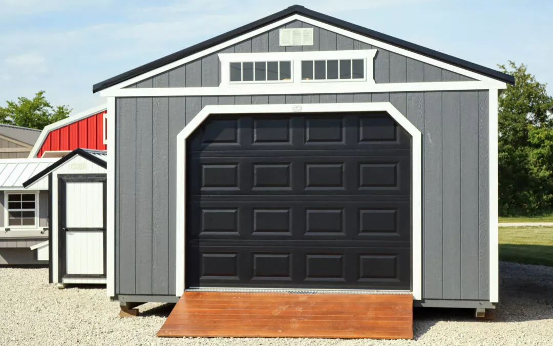 Cedar Ridge Sales Ramsey IL Ava IL A gray storage shed with a black garage-style door and a wooden ramp, situated on a gravel lot. Smaller sheds are visible in the background.