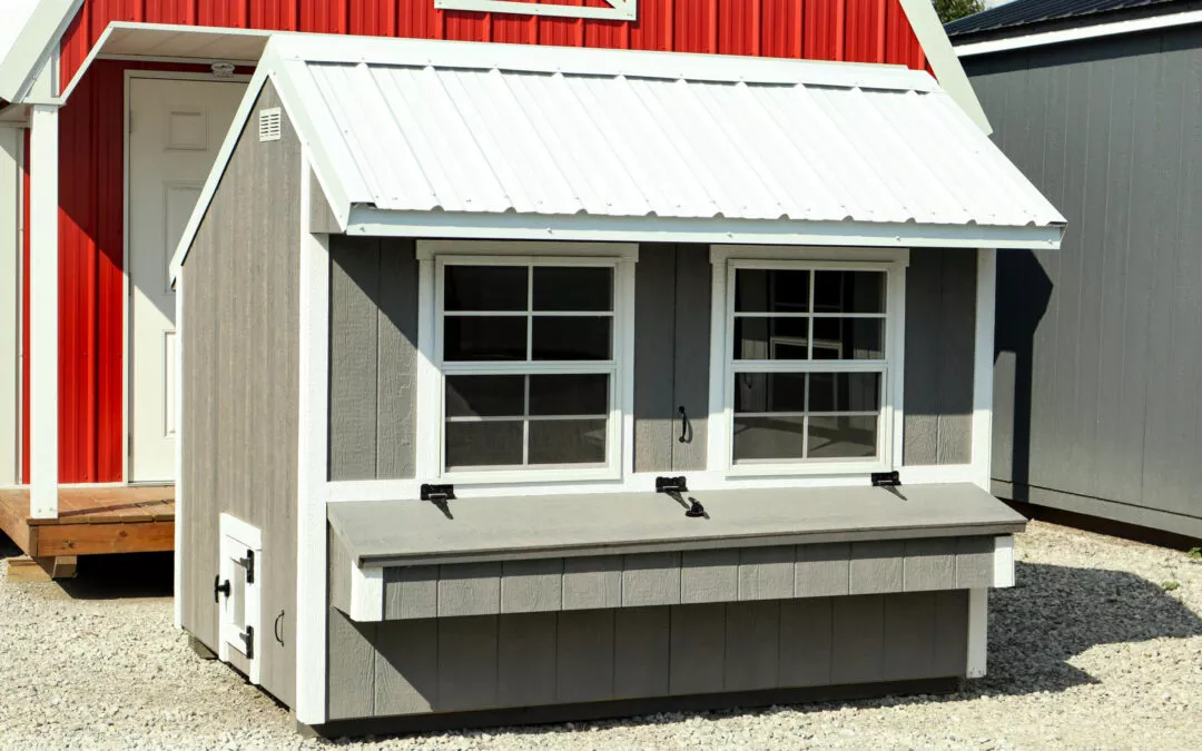 Cedar Ridge Sales Ramsey IL Ava IL A small grey and white shed with two front windows, located on a gravel surface, situated in front of a larger red and white building.