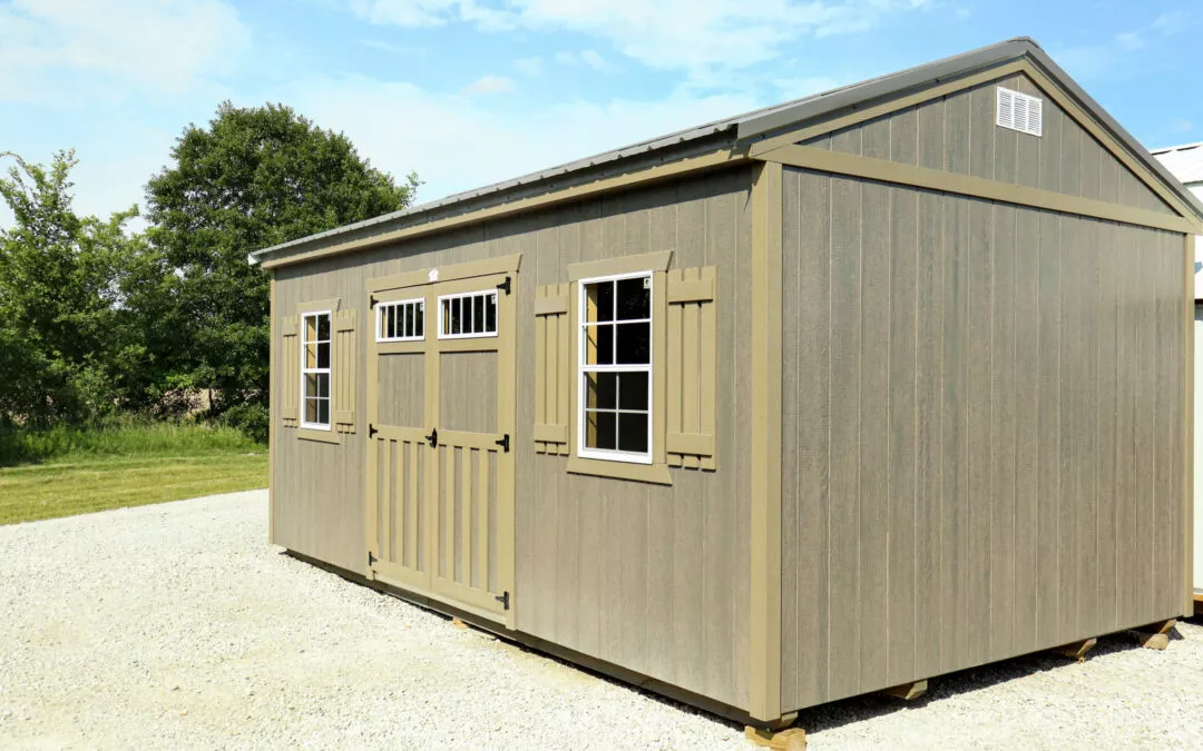 Cedar Ridge Sales Ramsey IL Ava IL A large, light gray wooden shed with double doors and windows, situated on a gravel surface with green trees and a blue sky in the background.