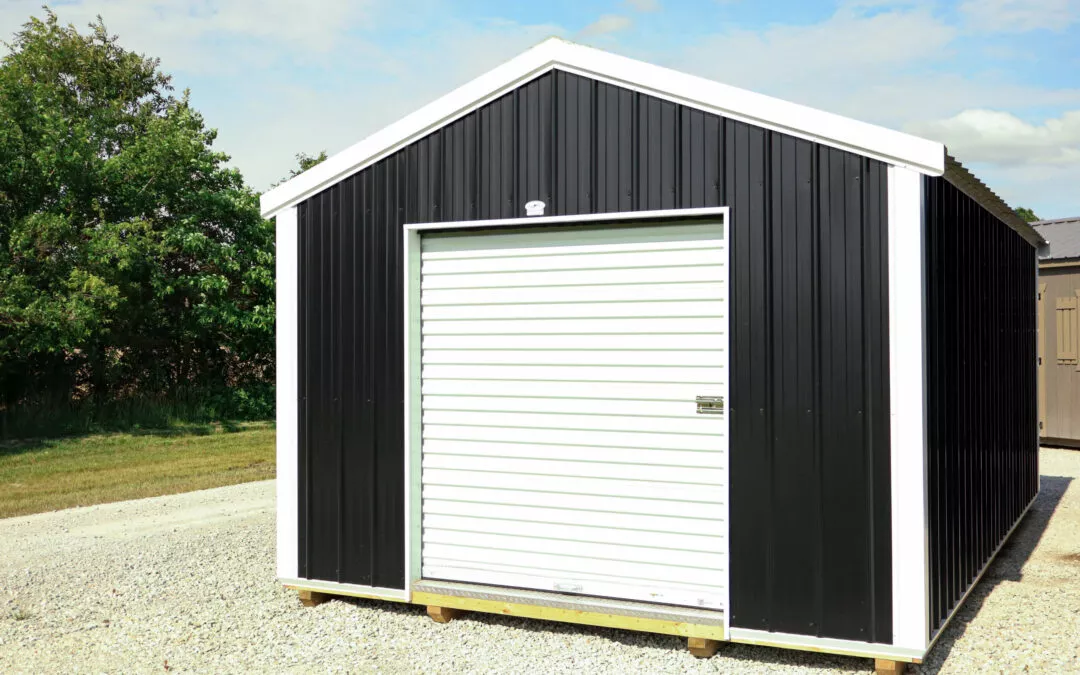 8.18.24 Cedar Ridge Sales Gallery 9 A small black and white shed with a closed roll-up door stands on a gravel surface next to a grassy area with trees in the background.