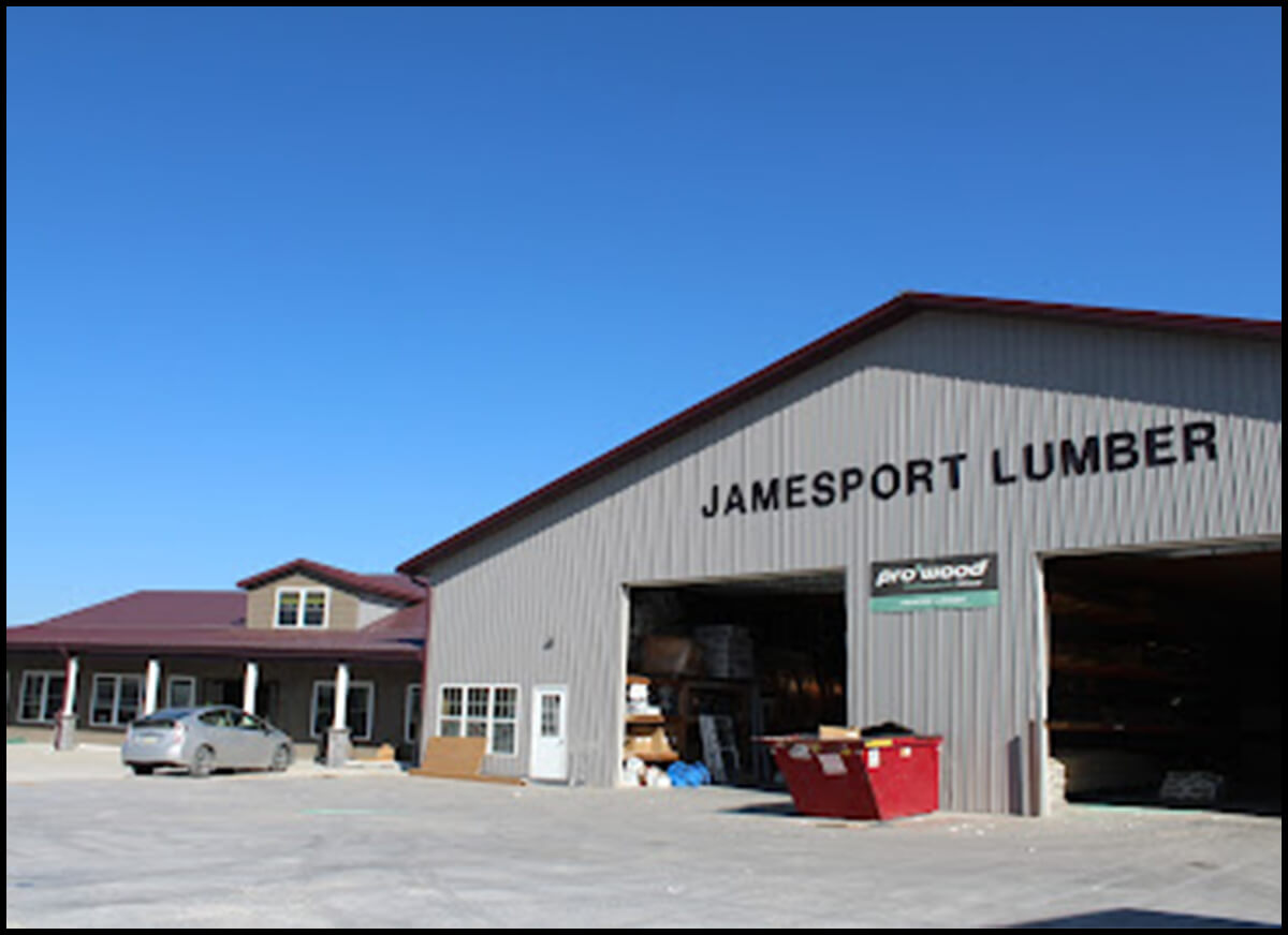 A gray metal building with red trim labeled "Jamesport Lumber." An open garage door reveals stacks of lumber. A parked car and a red dumpster are visible in front. Clear blue sky overhead.