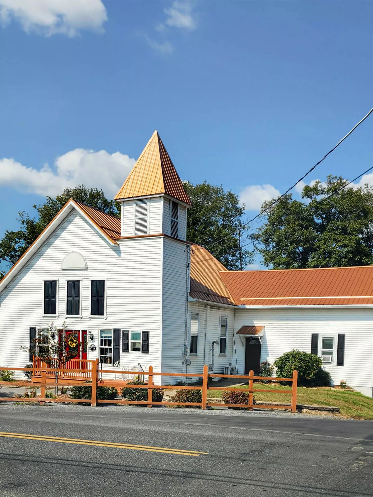 PREMEX Metal Roofing, Siding, & Trim Mifflinburg, PA A white church with a steeple and brown metal roof stands under a clear sky, with a small garden and wooden fence in front.