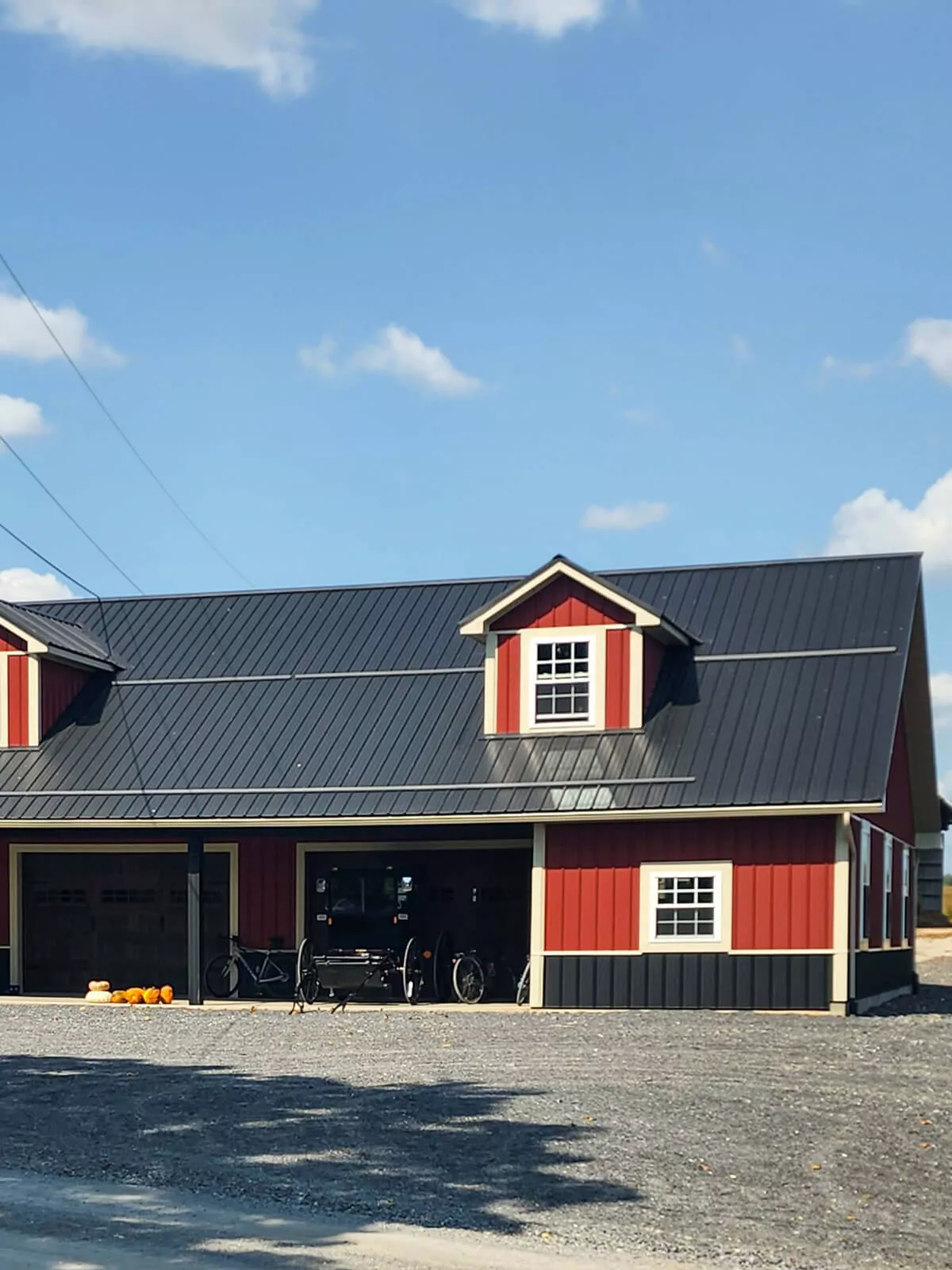 PREMEX Metal Roofing, Siding, & Trim Mifflinburg, PA A red and black barn with a metal roof and white trim featuring two small dormer windows. A horse cart and bicycle are parked outside, and several pumpkins are placed near the entrance.