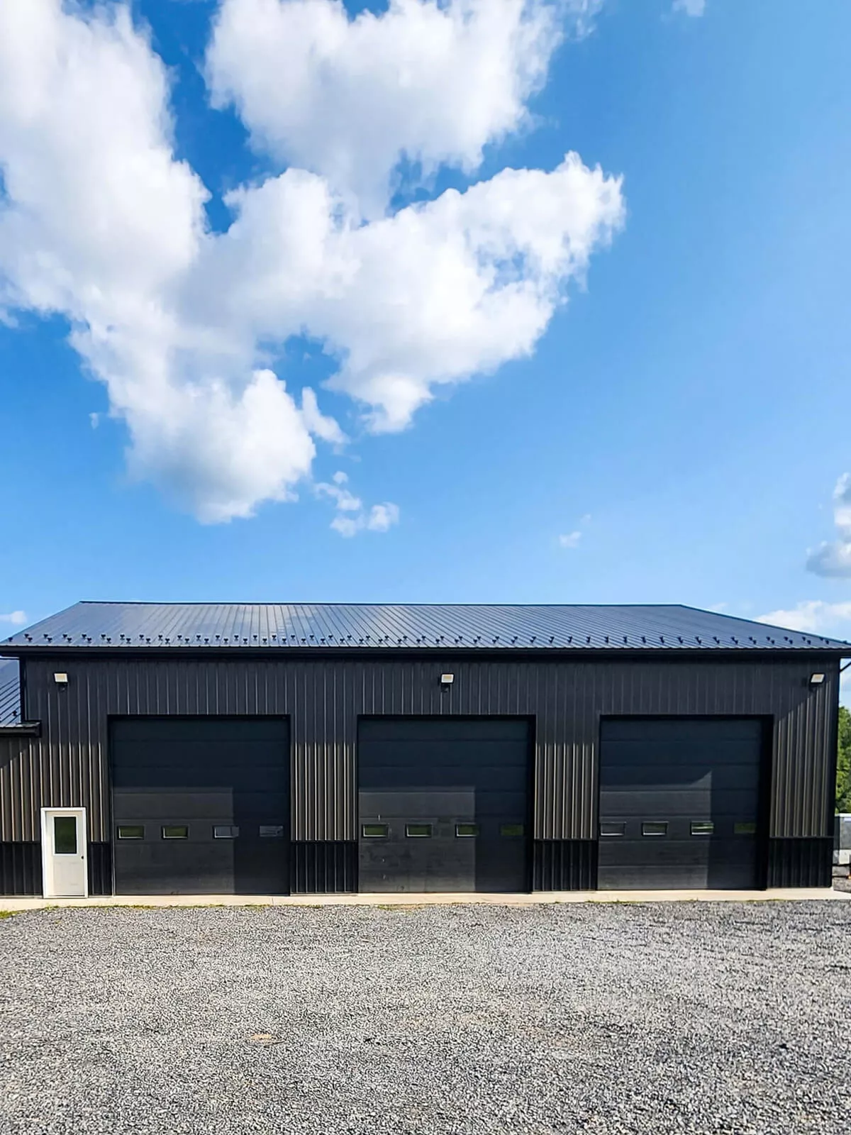 PREMEX Metal Roofing, Siding, & Trim Mifflinburg, PA A large, black industrial building with three closed garage doors and a metal roof under a clear blue sky with some clouds, stands on a gravel lot. A single white door is on the left side of the building.