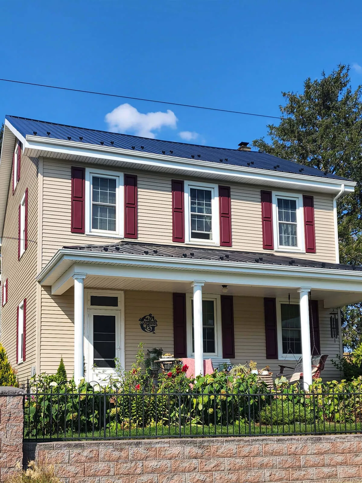 PREMEX Metal Roofing, Siding, & Trim Mifflinburg, PA Two-story house with yellow siding, white trim, a black metal roof, and maroon shutters. It has a covered front porch, a black metal roof, and a small garden in front, enclosed by a black metal fence.