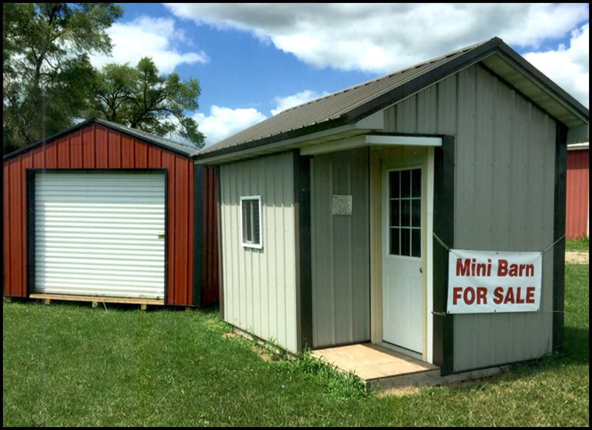 Zimmerman Construction & Mini Barns Sheridan, MI A small beige shed with a sign reading "Mini Barn For Sale" beside a larger red shed with a white roll-up door, set on a grass lawn under a partly cloudy sky.