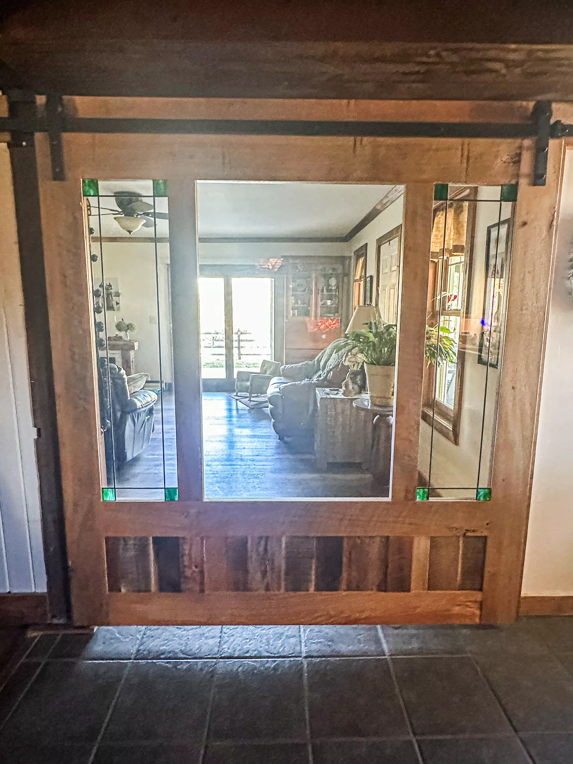 10.29.24 Cross Timbers Woodworking Review Ad A wooden sliding door with clear and green stained glass panels opens to a cozy living room with plants and a couch.