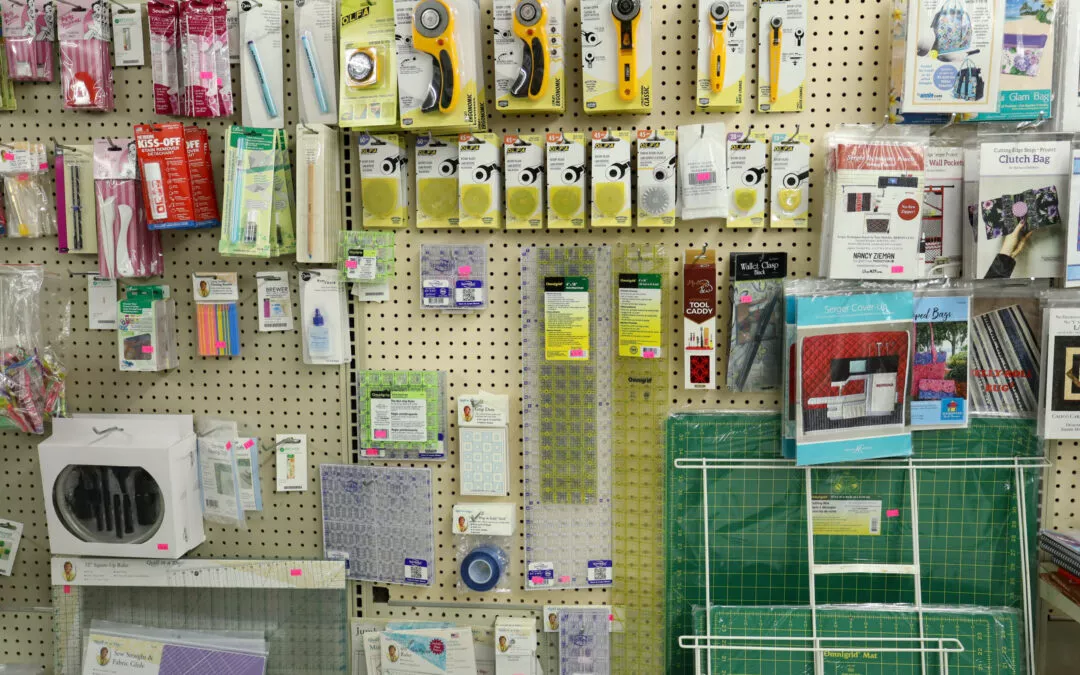 Brubaker's Sewing & Furniture Withee, WI A store display featuring various quilting and sewing supplies, including rotary cutters, cutting mats, rulers, fabric markers, needles, and pattern booklets, arranged on a pegboard wall.