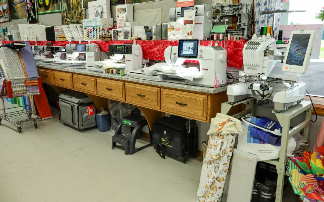 Brubaker's Sewing & Furniture Withee, WI A row of sewing and embroidery machines on a wooden counter in a craft store, with fabric and thread supplies nearby.