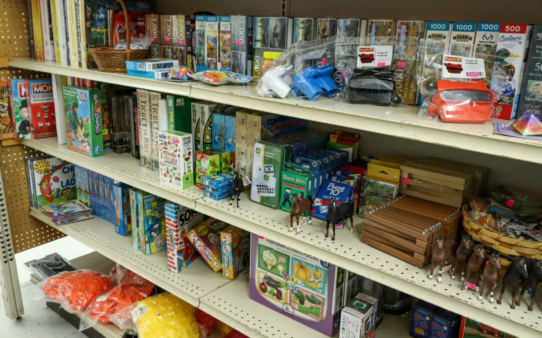 Brubaker's Sewing & Furniture Withee, WI Shelves in a store displaying various board games, puzzles, and toys including horses and LEGO sets. Items are neatly arranged and some are in plastic bags with price tags.