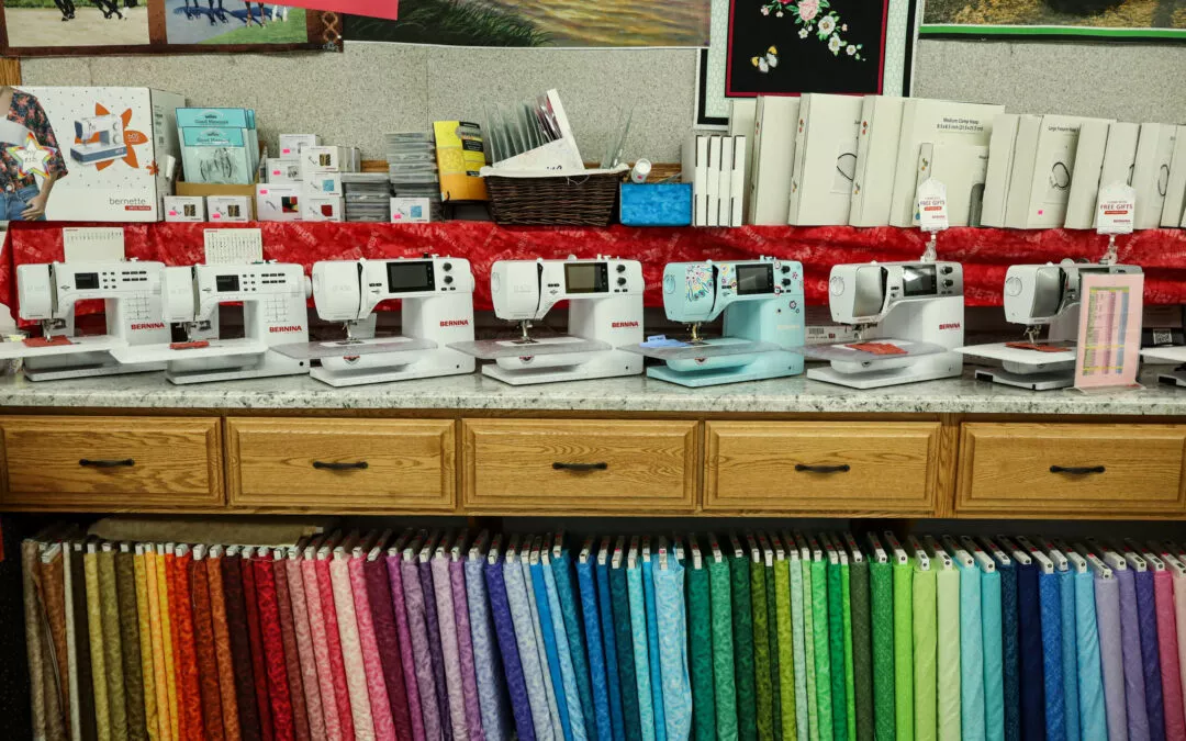 Brubaker's Sewing & Furniture Withee, WI A row of sewing machines displayed on a counter above a selection of colorful fabric bolts in a store.
