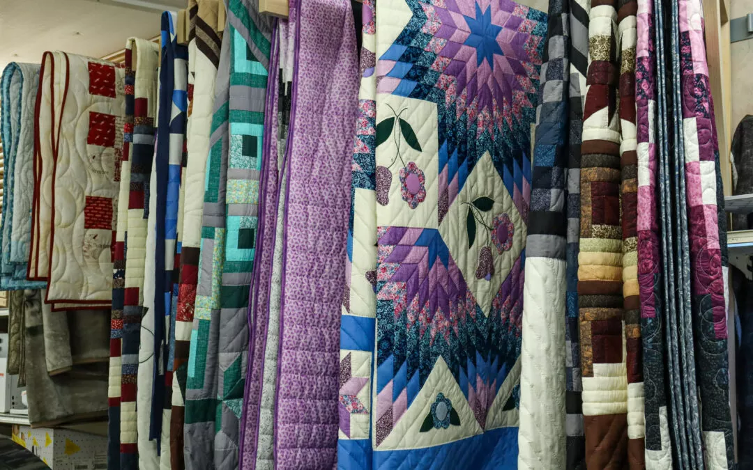 Brubaker's Sewing & Furniture Withee, WI A display of various colorful quilts hanging vertically, showcasing diverse patterns and designs.