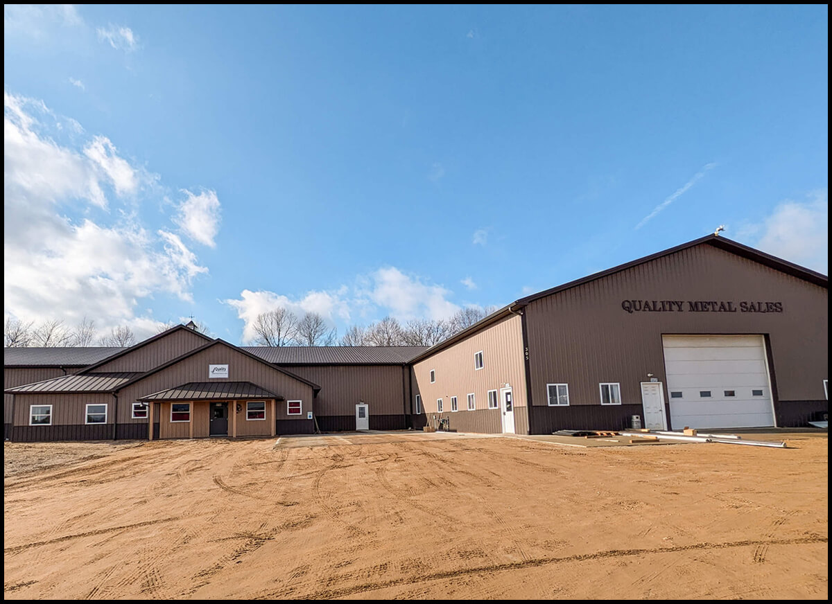 Quality Metal Sales Vermontville, MI Large brown warehouse labeled "Quality Metal Sales" with two buildings connected. Blue sky with clouds above and a dirt lot in front.