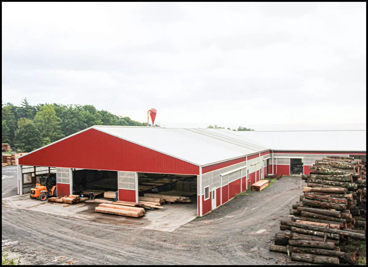 Yoder Lumber Co McAlisterville, PA A red and white industrial building with stacks of logs and wooden planks outside, surrounded by trees. An orange vehicle is parked nearby.