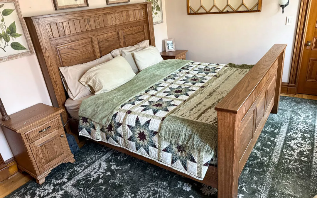 Sugar Valley Chair Shop Loganton, PA Bedroom with a wooden bed frame, patterned quilt, and green throw blanket. Nightstands and framed botanical prints adorn the walls. A patterned rug covers the floor.