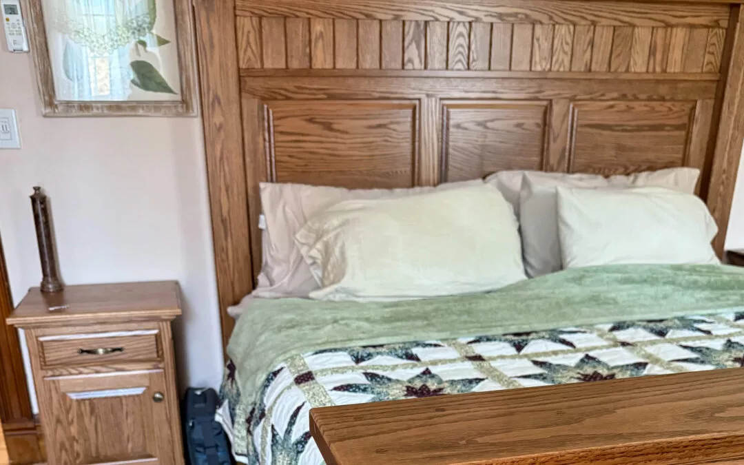 Sugar Valley Chair Shop Loganton, PA A neatly made bed with a wooden headboard, two pillows, and a patterned quilt. A wooden nightstand with a drawer sits beside the bed. Wall artwork is visible in the background.