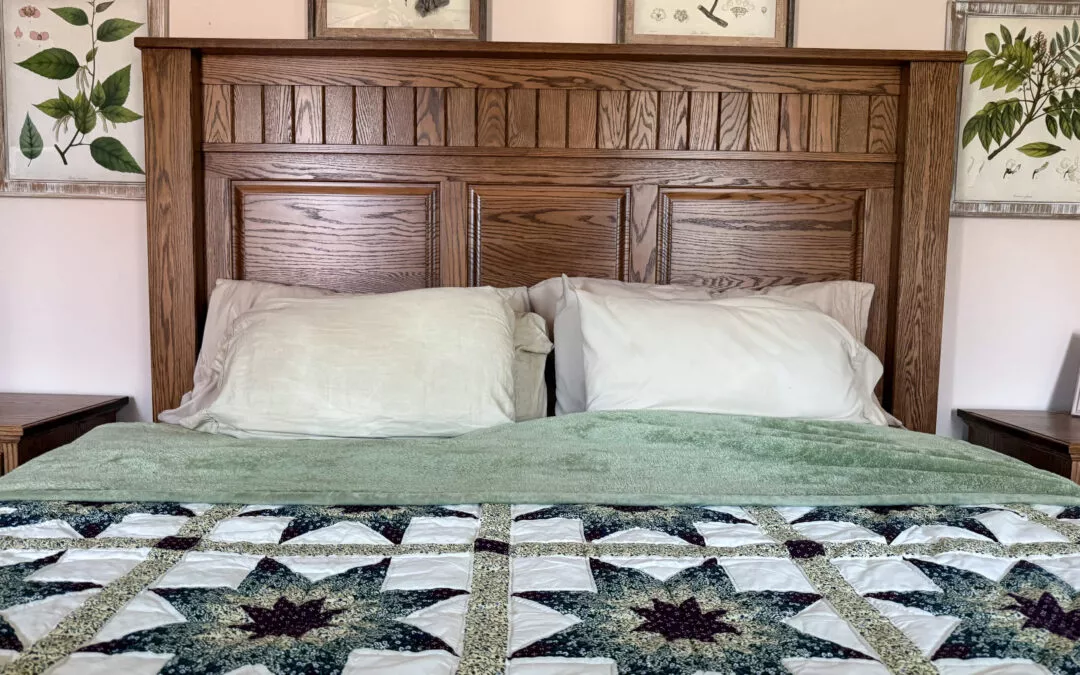 Sugar Valley Chair Shop Loganton, PA A neatly made bed with a wooden headboard, green blanket, and decorative quilt, flanked by two nightstands. Botanical prints are displayed on the wall above.