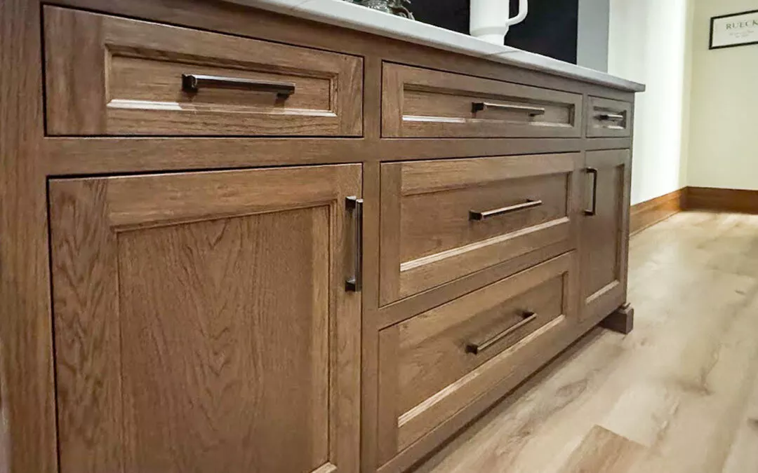 The Woodpecker Sullivan, Illinois Wooden kitchen cabinets with black handles, featuring multiple drawers and cupboards, set on a light wood floor. A countertop with a plant and a white pitcher is visible above.