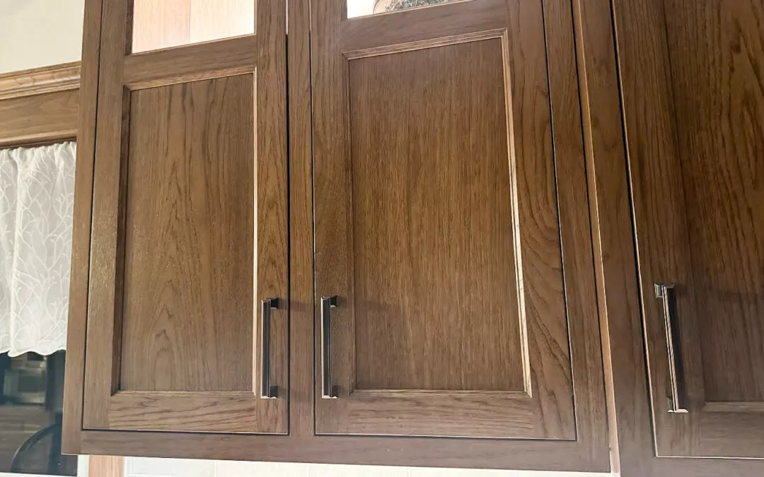 The Woodpecker Sullivan, Illinois Wooden kitchen cabinets with metal handles, positioned above a light-colored tiled backsplash and framed by a window with a lace curtain.