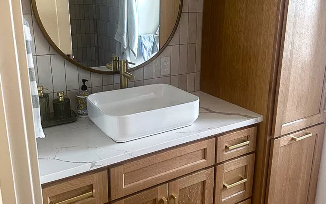 The Woodpecker Sullivan, Illinois Bathroom vanity with wooden cabinets, a round mirror, square white sink, gold faucet, and toiletries.