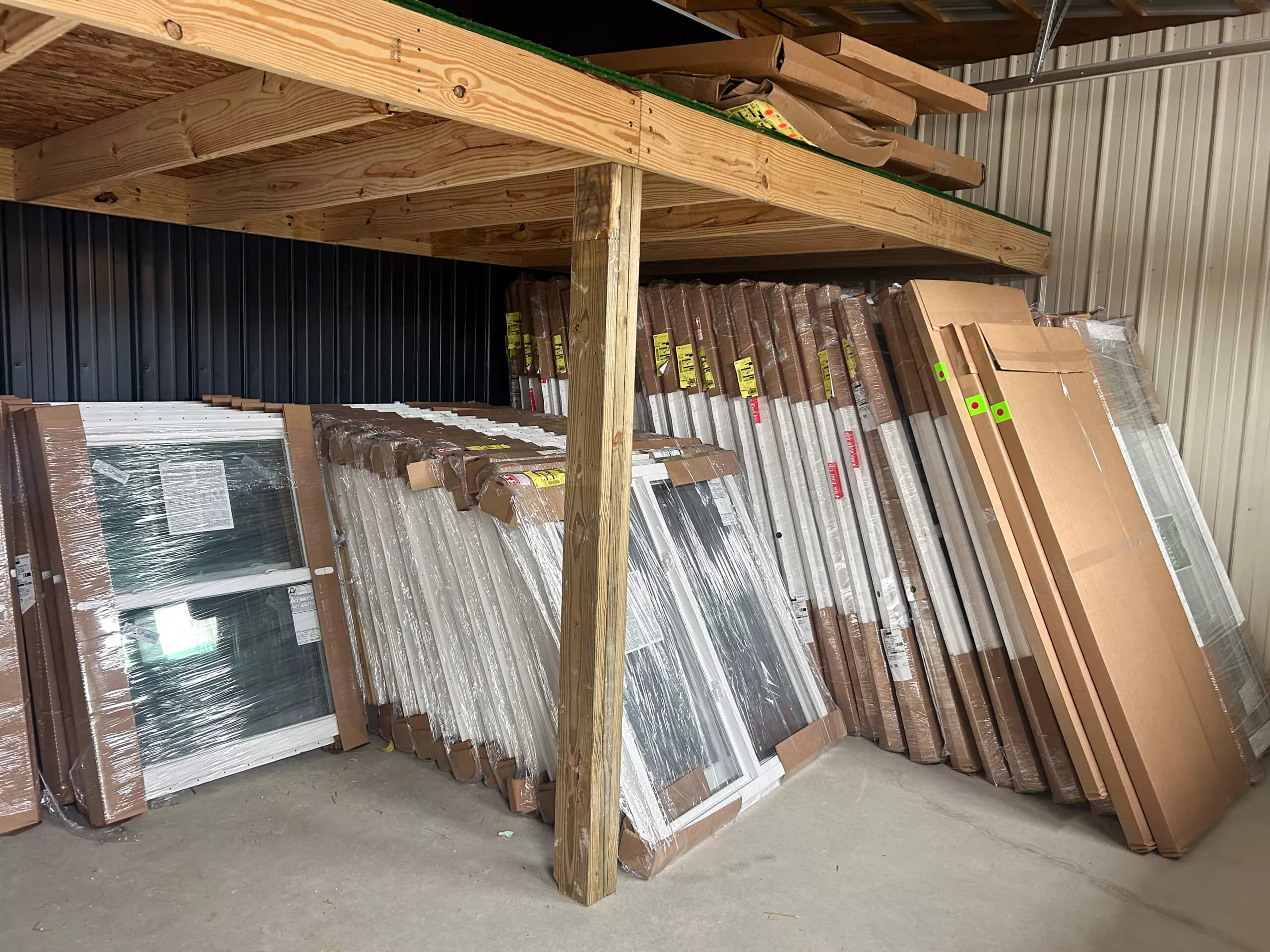 Amish Ridge Metals Perry, OH Stacks of windows and doors wrapped in plastic and cardboard are stored upright against a wall under a wooden mezzanine in a warehouse.