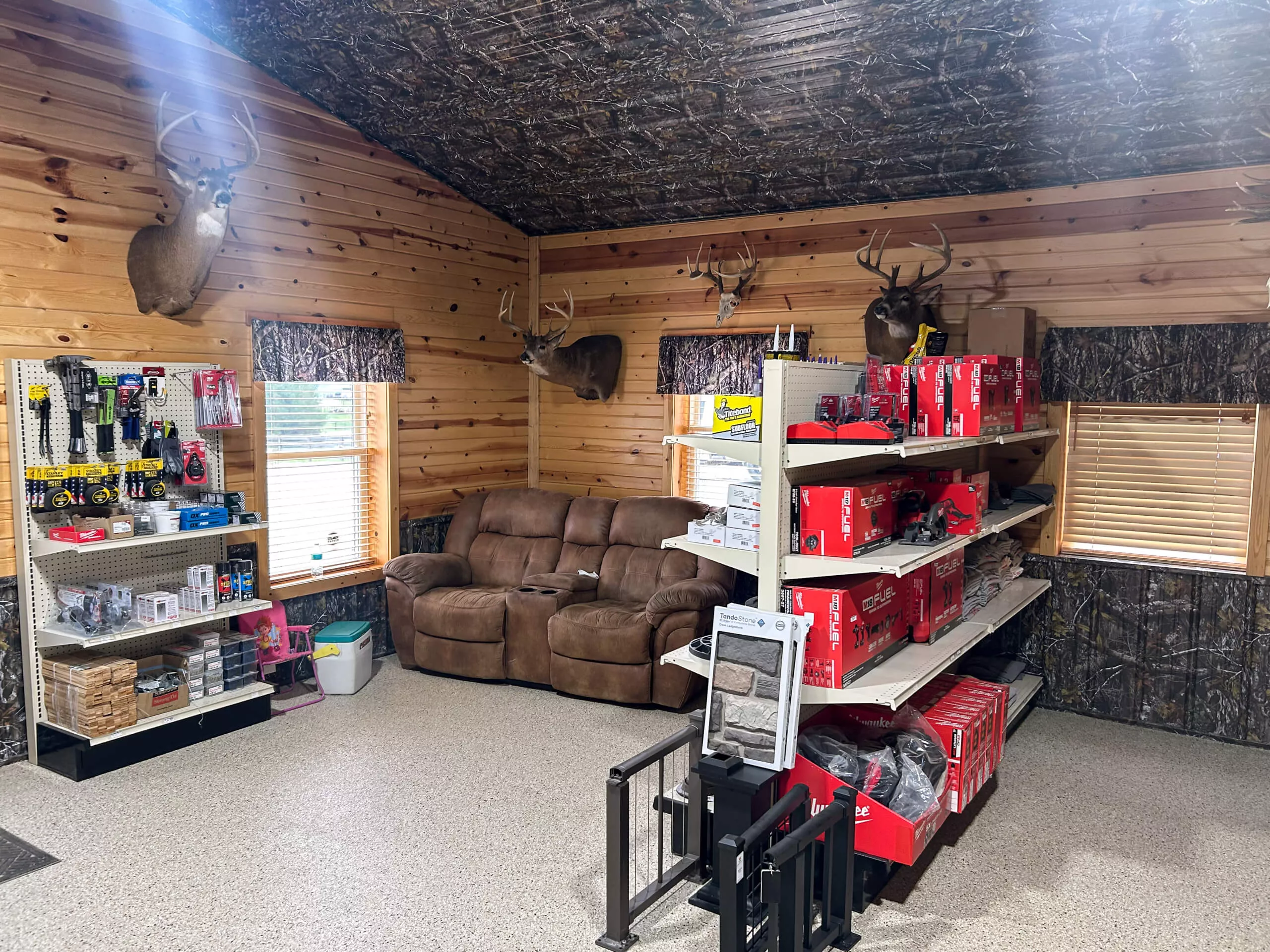 Amish Ridge Metals Perry, OH Interior of a store with wooden walls, deer mounts, a brown couch, shelves with red-boxed products, and a display of small packaged items. Light from two windows brightens the space.