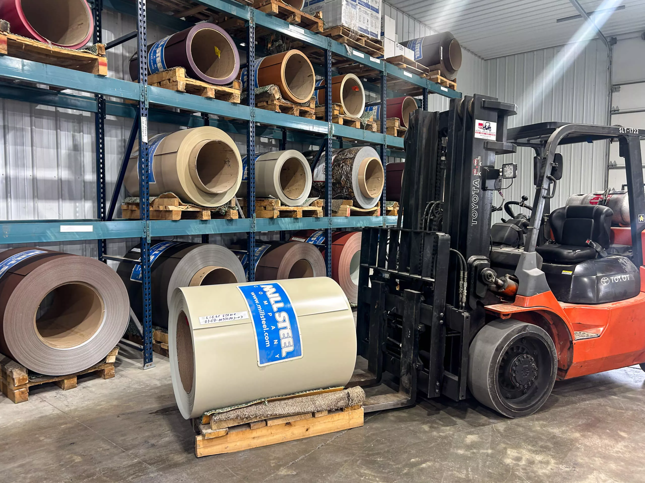 Amish Ridge Metals Perry, OH A forklift moves a large roll of steel labeled "All Steel" in a warehouse with multiple steel coils stored on shelves.